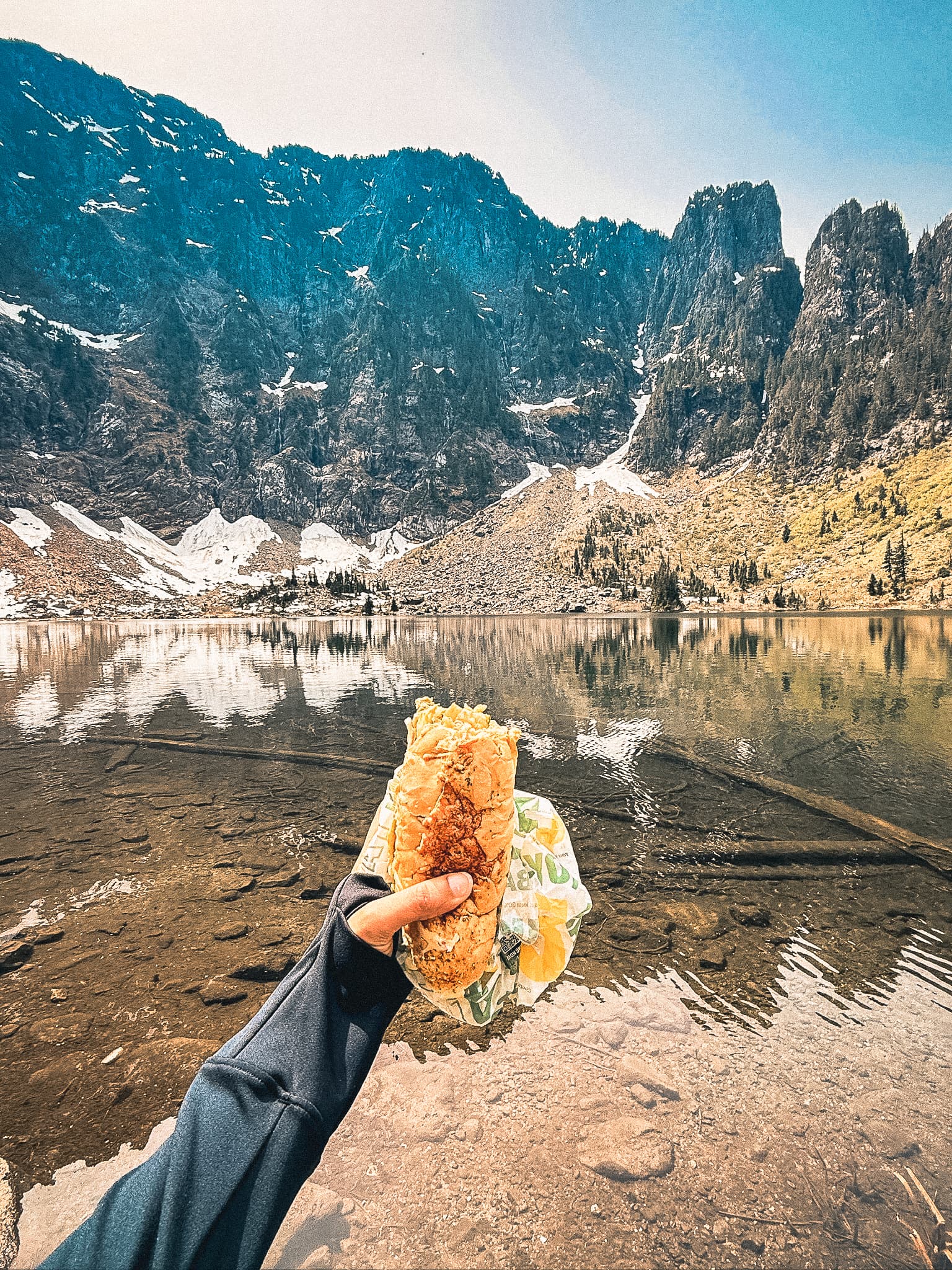 Hand holding a burrito in front of Lake 22 with snow-capped peaks behind
